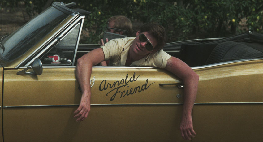 A man with muscular arms and sunglasses leans over the door of a gold convertible. His head is tipped to one side. The name Arnold Friend is painted in script letters on the side of the car.