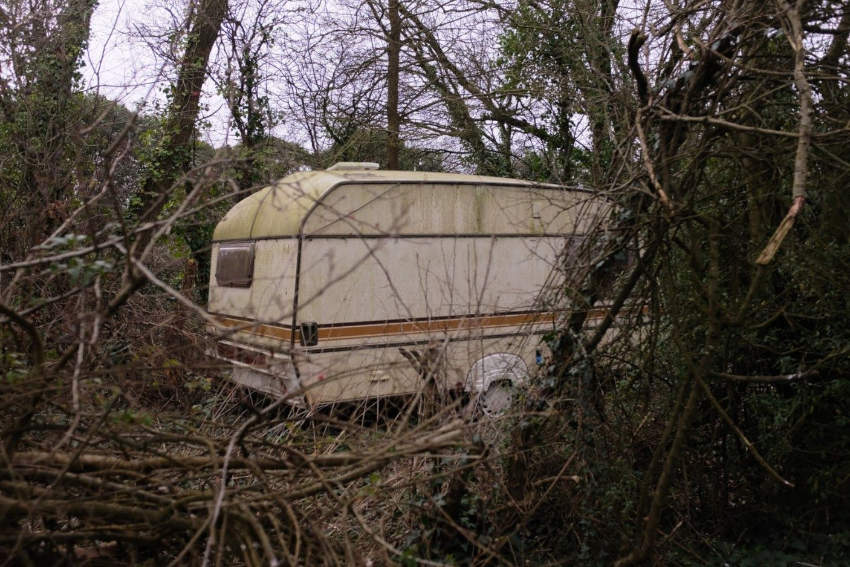 A caravan parked in the corner of a field surrounded by trees and undergrowth.