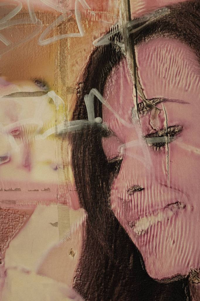 A close up of a shop fascia which has warped and cracked in the sun so that the woman on the image has blank, black eyes and a distorted, mad smile.