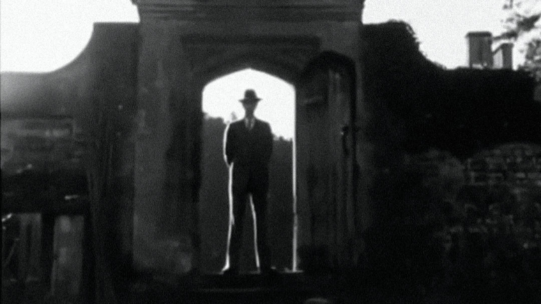 A creepy man in a hat framed in a stone archway.