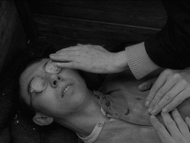A young man with shattered spectacles lies on the ground while an old woman's hand reaches out for his face.