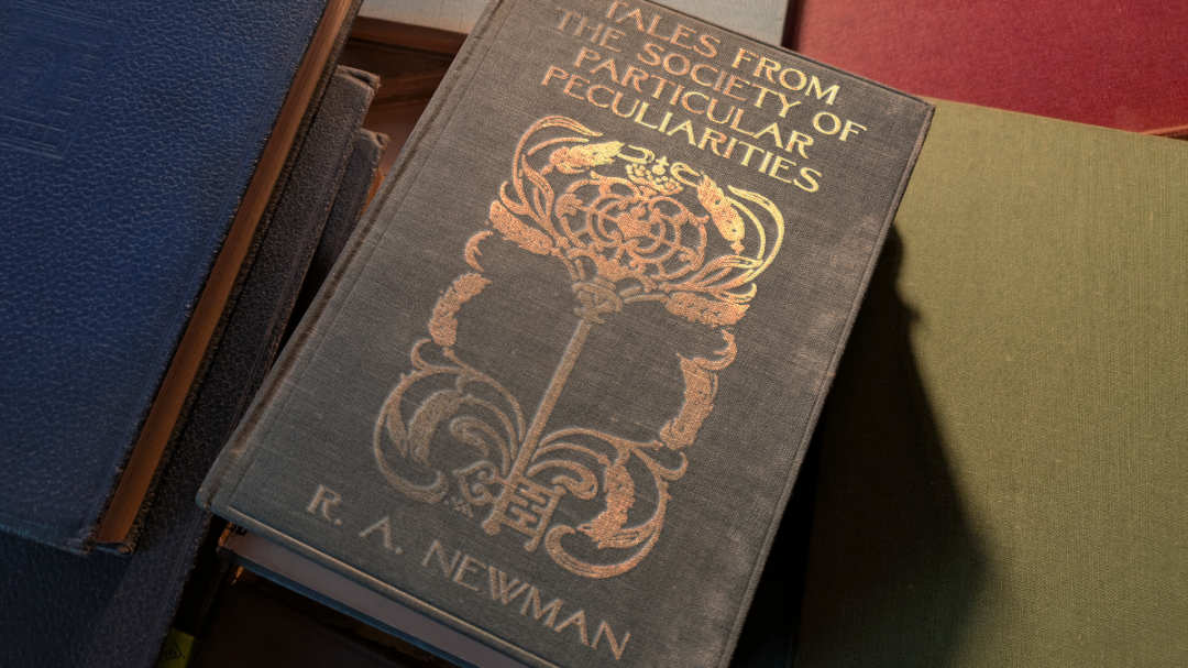 A stack of books including what looks like an Edwardian hardback with Art Nouveau design: Tales from the Society of Peculiar Particularities.