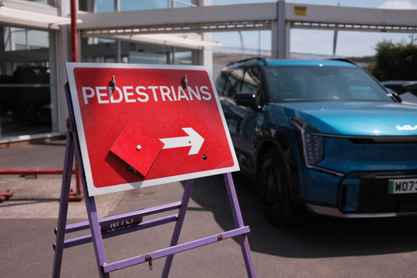 A temporary sign outside a car dealership directs pedestrians around road works. A big blue car is in the background.