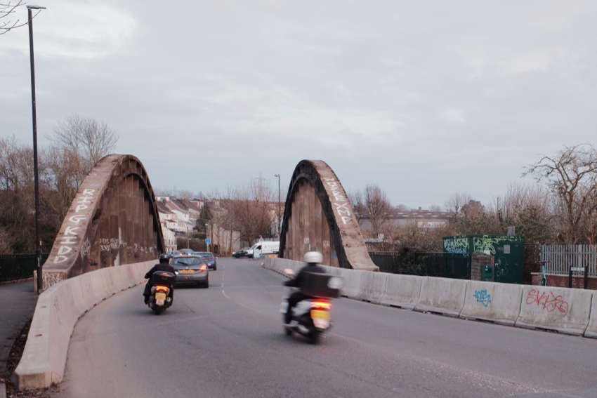 A concrete bridge with two big arches suspending it. There is graffiti on the concrete. Mopeds are speeding along the road, blurred.