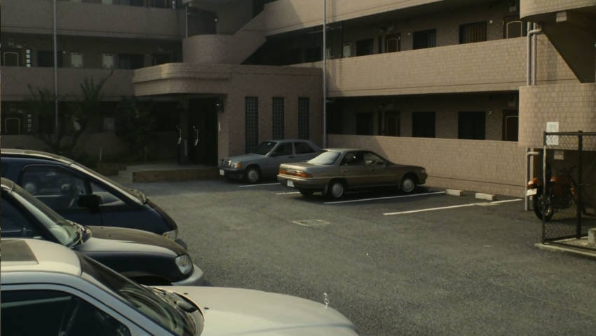 A bland car park behind a bland apartment block with several white, grey and beige cars.