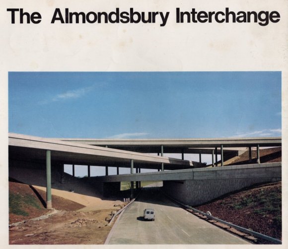 A 1960s photo of a mini driving along an empty road towards multi-layered motorway flyovers. The sky behind is very blue.