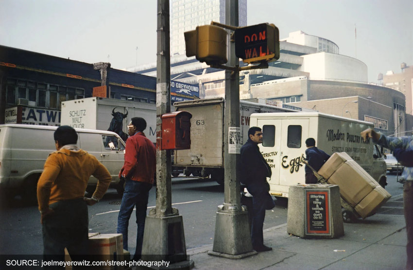 A group of men gather on a pavement on a busy street in an American city. One wears a yellow top, another a red jacket. There is a yellow and red traffic signal next to them.
