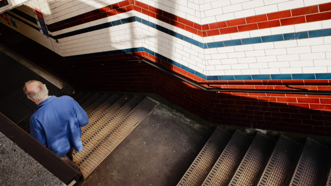 A man in a blue jacket disappearing into a tube station entrance with filthy steps and diagonal lines in various colours on the wall.