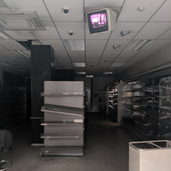 An empty, abandoned shop with bare shelves, dark shadows, and security monitor on the ceiling glowing purple.