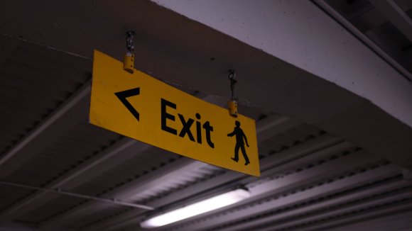 A dangling yellow exit sign in a brutalist car park with poor lighting.