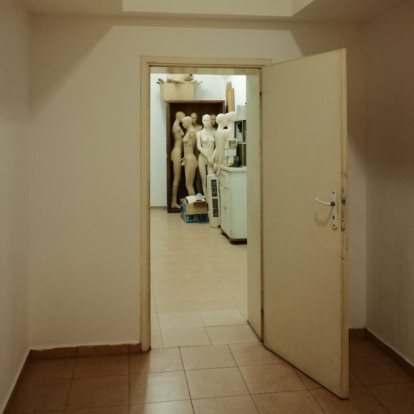 A door in a dingy basement through which can be seen a group of creepy white mannequins gathered around a wardrobe.