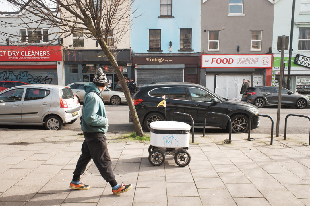 A delivery robot on a city street being followed and observed by a pedestrian.